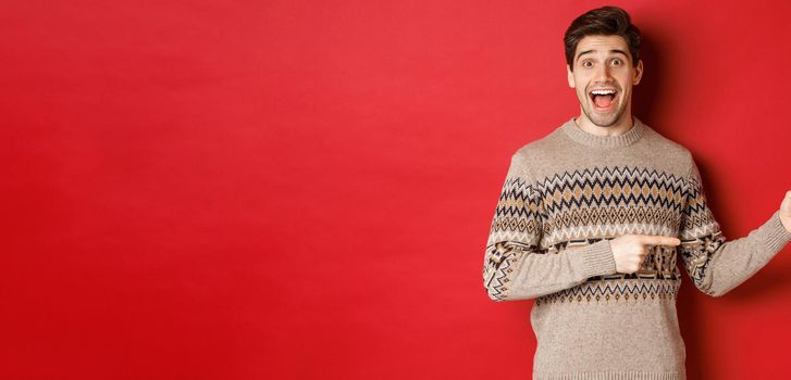 Concept of christmas celebration, winter holidays and lifestyle. Handsome man in xmas sweater pointing fingers right, smiling amazed, showing new year promo against red background.