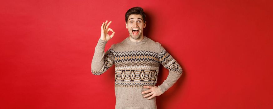 Concept of christmas celebration, winter holidays and lifestyle. Amazed and happy man in xmas sweater, showing okay sign and looking at something fantastic, red background.