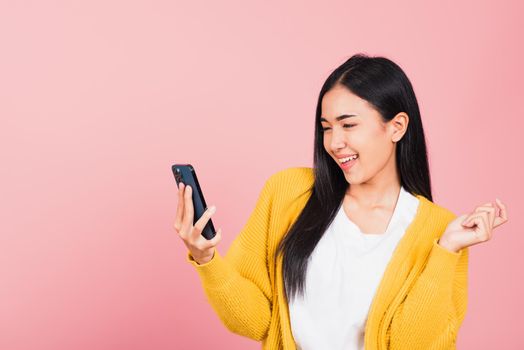 Happy Asian portrait beautiful cute young woman excited laughing holding mobile phone, studio shot isolated on pink background, female using funny smartphone making winner gesture