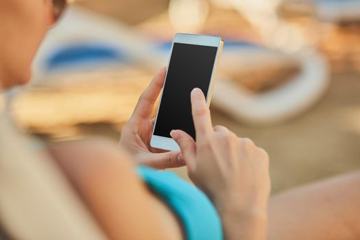 Close up view of a young attractive woman on holiday laying down on a white chaise-longue, holding and using a smartphone. Black display. People travel technology.