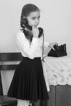 A beautiful little schoolgirl girl in a white blouse and black long skirt, with neatly braided pigtails on her head.She is talking on an old phone. Interior of the fifties of the last century.