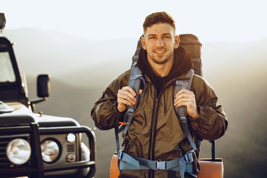Portrait of a young traveler man in hiking equipment standing near his off-road car at dawn