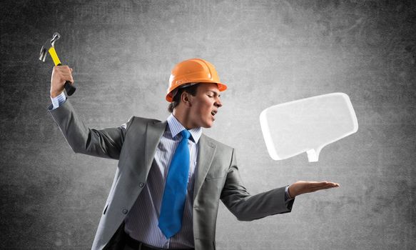 Furious businessman going to crash glass speech bubble with hammer. Young handsome man in business suit and safety helmet standing on wall background. Business communication and online dialogue