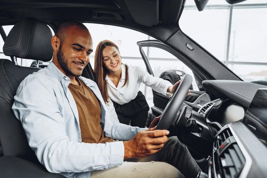 Young attractive caucasian woman salesperson in car showroom showing a car to her male African American client