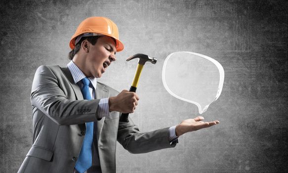 Furious businessman going to crash glass speech bubble with hammer. Young handsome man in business suit and safety helmet standing on wall background. Business communication and online dialogue
