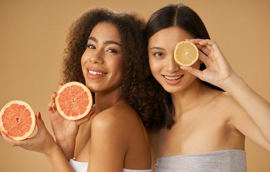 Portrait of two pretty mixed race young women holding grapefruit and lemon cut in half while posing together isolated over beige background. Health and beauty concept