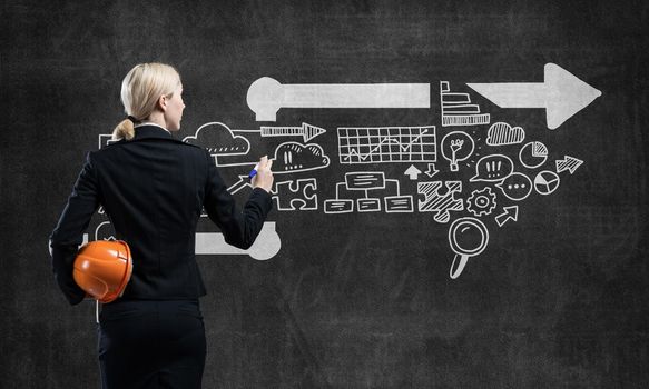 Businesswoman draws arrows consists of business doodles on blackboard. Back view woman with safety helmet and marker pen in hands. Idea generation and business development. Coaching and mentoring.