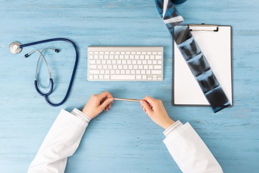 Top view of doctor hands holding pen. X-Ray radiography examination in hospital. Physician sitting at blue wooden desk. Examination and consultation in clinic. Workplace with stethoscope and keyboard.