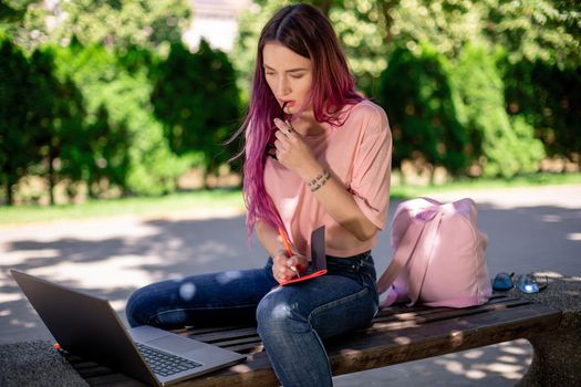 Woman writing in a notebook sitting on a wooden bench in the park. Girl working outdoors on portable computer, copy space. Technology, communication, freelance and remote working concept.