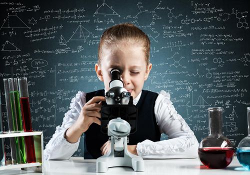 Little girl scientist looking through microscope in classroom at chemistry lesson. School chemical laboratory with glass flasks and test tubes. Schoolgirl making experiment in chemistry class.