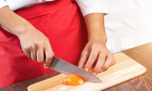Chef hands cutting orange fresh pepper on wooden board. Close up man hands cooking with knife on table in kitchen. Professional chef in red apron on light background. Restaurant food preparation.