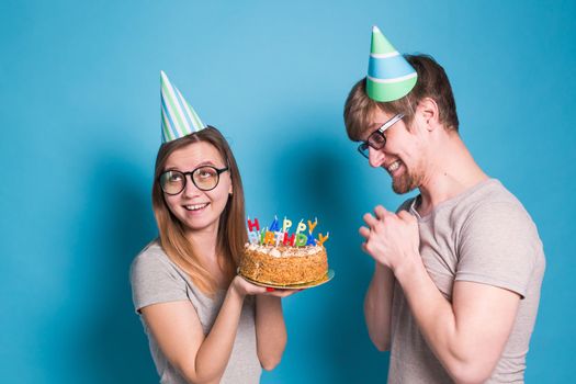 Funny young girl congratulates her boyfriend happy birthday holding a cake with candles in her hands standing on a blue background. Concept of congratulations and warm relations