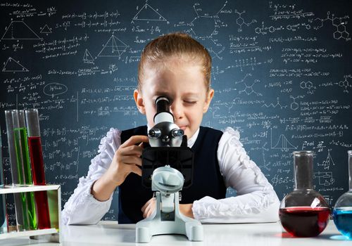 Little girl scientist looking through microscope in classroom at chemistry lesson. School chemical laboratory with glass flasks and test tubes. Schoolgirl making experiment in chemistry class.