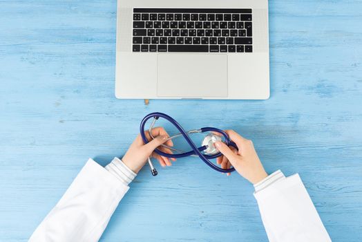 Top view of doctor hands with stethoscope. Therapist in white coat sitting at blue wooden desk with laptop. Examination and consultation in clinic. Medical application and online healthcare services.