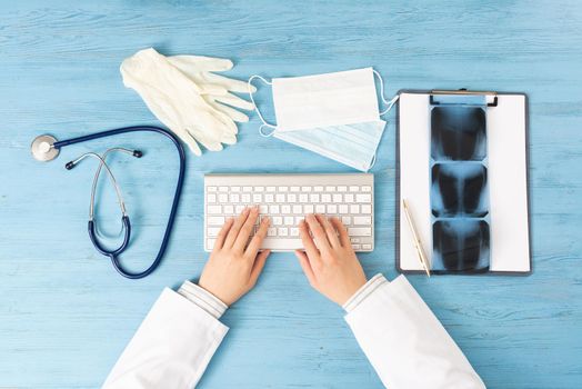 Top view of doctor hand typing at keyboard. Radiography examination in hospital. Therapist sitting at blue wooden desk with stethoscope, medical gloves and mask. Examination and consultation in clinic