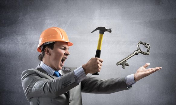 Furious businessman going to crash with hammer steel key. Young handsome contractor in business suit and safety helmet standing on grey wall background. Financial crisis in real estate. Password hack