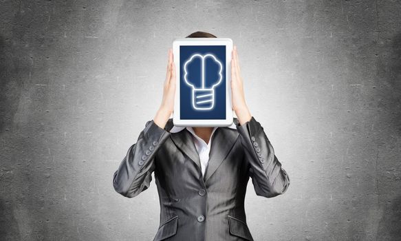 Portrait of woman covering her face with tablet computer. Businesswoman showing tablet PC with idea lightbulb sign on screen. Corporate businessperson on grey wall background. Digital technology