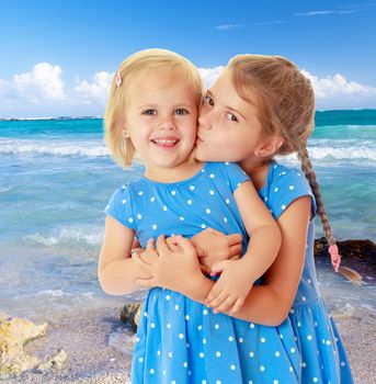 Two charming little girls, sisters , in identical blue dresses with polka dots. Older sister kissing the younger on the cheek.On the background of blue sea and blue sky with clouds.