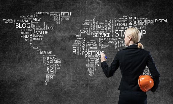 Businesswoman standing near world map on concrete wall. International company business presentation. World communication and globalization. Back view woman with safety helmet and marker pen in hands.