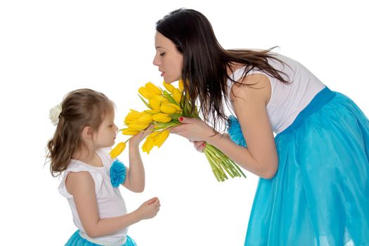 Beautiful young mother and little daughter in the same blue long skirts tutus , standing sideways to the camera ,admiring the bouquet of yellow tulips.Little girl enjoying the smell of flowers.Isolated on white background.