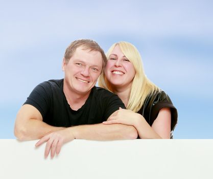 A man and a woman tenderly hugging each other. Peeping over white banner.On the background of summer blue sky and fluffy clouds.