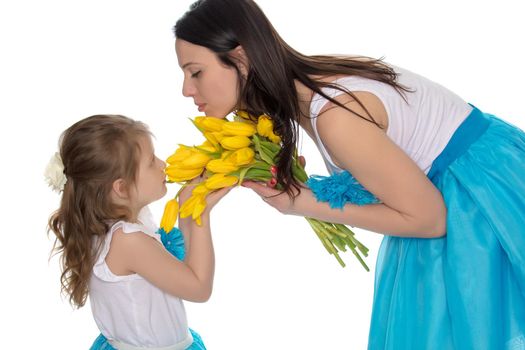 Beautiful young mother and little daughter in the same blue long skirts tutus , standing sideways to the camera ,admiring the bouquet of yellow tulips.Little girl enjoying the smell of flowers.Isolated on white background.