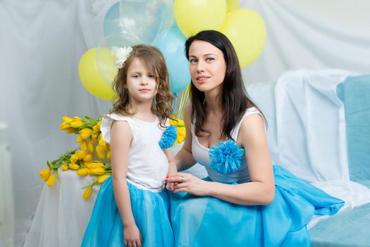 Beautiful young mother and little daughter in the same blue dresses on the sofa with a bouquet of flowers.