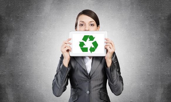 Businesswoman holding tablet computer with recycle sign on screen. Beautiful woman in business suit show tablet PC near her face. Corporate businessperson on grey wall background. Digital technology