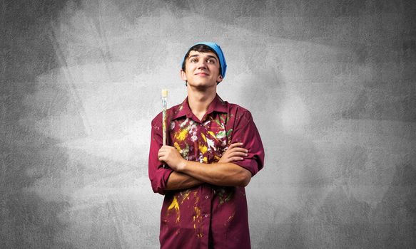 Pensive painter artist standing with folded arms on gray wall background. Portrait of young man looking up dreamily. Artistic occupation and creative profession. Art school student posing.