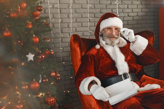Santa Claus having a rest by the Christmass tree. Home decoration. Santa with letter from children.