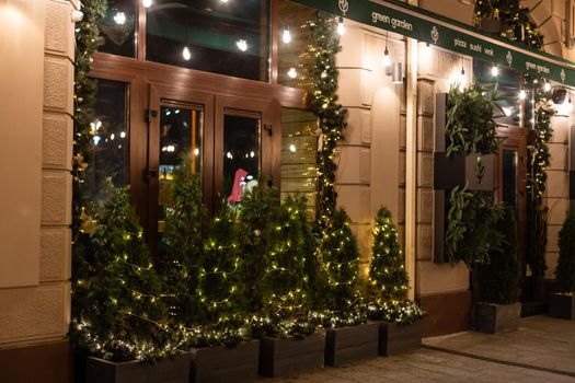 Stylish christmas tree with red berries, wreath on doors and wooden sleigh at front of store at holiday market in city street. Christmas street decor. Space for text. Rustic decoration.