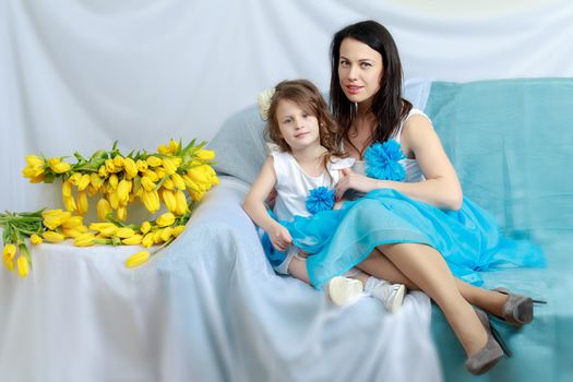 Beautiful young mother and little daughter in the same blue dresses on the sofa with a bouquet of flowers.