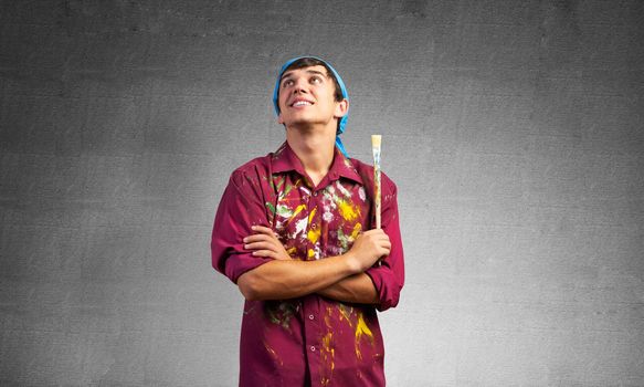 Smiling young painter artist with folded arms. Portrait of happy decorator looking upwards on gray wall background. Artistic occupation and creative profession. Art school student posing.
