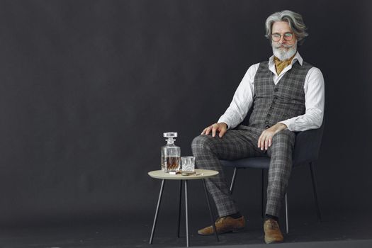 Close up portrait of grinning old-fashioned man. Elegant man in a studio. Gentelman sitting on a chair. Grandfather with a glass of whiskey.