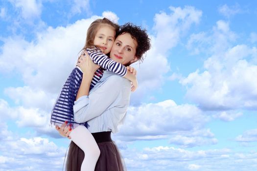 Happy young beautiful mother,lifted his little beloved daughter .And the girl embraced her mother's neck and pressed my face to her cheek.On the background of summer blue sky and fluffy clouds.