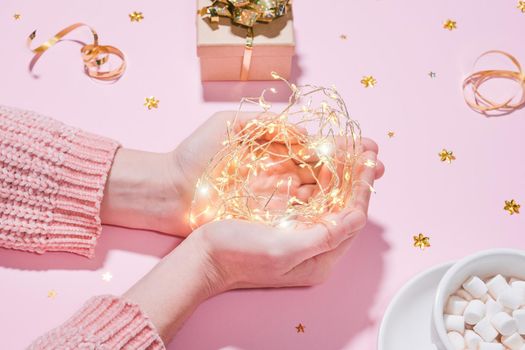 Beautiful and cozy holiday concept Hands holding string garland lights, on the background of wrapped gift, a cup of hot chocolate or cocoa with marshmallows, golden tinsel isolated on pink background