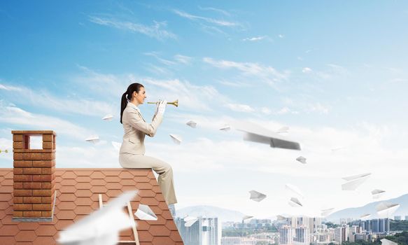 Attractive young woman playing music on roof of building. Brave businesswoman with trumpet instrument sitting on edge of roof. Flying paper planes in blue sky. Music performance on roof in modern city