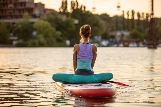 Young woman doing yoga on sup board with paddle. Yoga pose, side view - concept of harmony with the nature, free and healthy living, freelance, remote business.