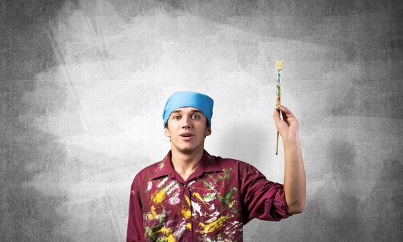 Young painter artist pointing upwards. Man holds paintbrush over his head. Portrait of caucasian painter on gray wall background. Creative hobby and artistic occupation. Art school student posing.