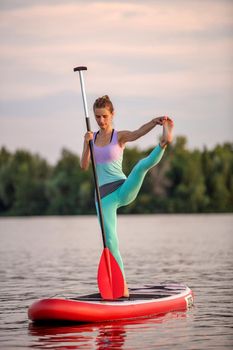 Sporty woman in yoga position on paddleboard, doing yoga on sup board, exercise for flexibility and stretching of muscles. Woman practicing yoga on the paddle board in the morning
