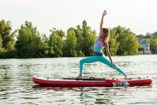 Young woman doing yoga on sup board with paddle. Yoga pose, side view - concept of harmony with the nature, free and healthy living, freelance, remote business.