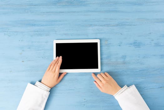 Top view of doctor hands with tablet computer. Therapist in white coat sitting at blue wooden desk. Examination and consultation in clinic. Medical application and online healthcare services.