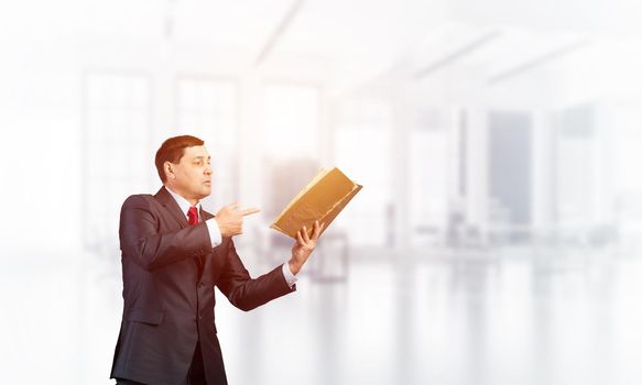 Senior businessman finger pointing into open book. Portrait of adult man in business suit and tie standing in blurred office interior. Manager holding big handbook. Professional reference information.
