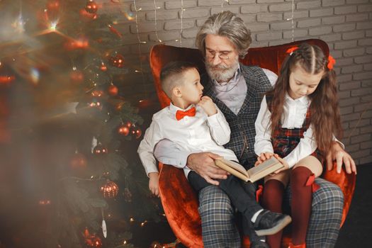 Grandfather wearing glasses, reading a book to small granddaughters twins in a room decorated for Christmas on the background of a Christmas tree. Christmas holiday concept. contrast photography