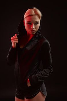 Beautiful seductive sporty girl with the slim sexy figure in the black hoodie, sport underwear and little black shorts is posing in the studio. Color filters.