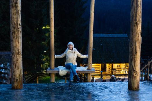 A woman in a down jacket with a hood on a swing in the city square. Evening background image of Christmas and New Year. Festive time.