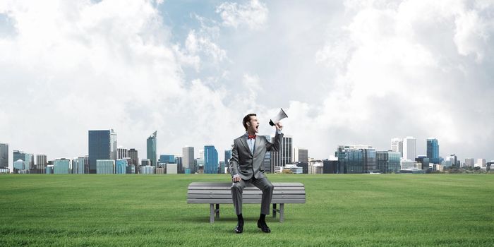 Young businessman sitting on bench and screaming emotionally in megaphone