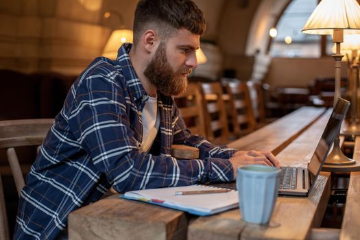 Young professional surfing the Internet on his laptop with cup of coffee on table at coffee shop or home office, working from cafe concept