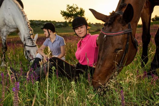 Friendly women are sitting next to a brown and white horses, which grazed in the meadow. Riders and horses relaxing outdoor in summer time.