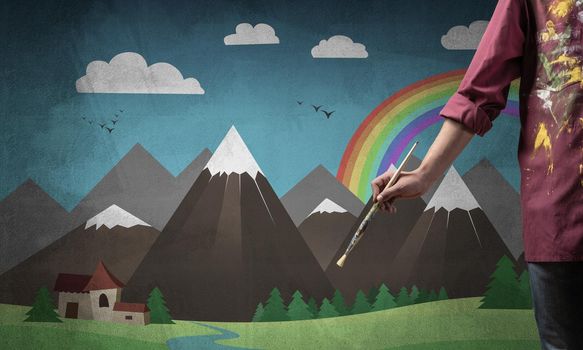 Close up artist hand holding paintbrush. Painter in shirt standing on background colorful picture. Summer landscape with mountains, blue sky and rainbow artwork. Creative hobby and profession.